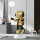 The Celestial Gold Astronaut Glow Balloon Sculpture by Giant Sculptures, standing on a white circular base with headphones and clutching a glowing balloon, beautifully complements the modern rooms grey walls and assorted decor, adding a unique space-themed charm with books and plants.