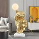 The contemporary living room features a 110cm Gilded Gold Astronaut Kitty Pup Glow Balloon Sculpture by Giant Sculptures, showcasing a gold astronaut with cat ears holding a polar bear on a white base. The space-themed decor is complemented by gray couches, wood panels, and yellow accents.