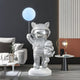 The Giant Sculptures Cosmic Chrome Astronaut Kitty Pup Glow Balloon Sculpture - 110cm adds a gleaming space-themed touch beside a modern gray sofa with cushions, perfectly complementing the futuristic setting that includes a small round side table, lamp, and potted plant against dark and light-colored walls.