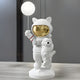 A Nova White Astronaut Kitty Pup Glow Balloon Sculpture by Giant Sculptures, featuring a bear in an astronaut suit with a glowing balloon and shiny gold visor, cradles an animal under one arm. The minimalist scene includes gray walls and vertical lines, enhancing its whimsical charm.