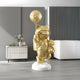 A majestic Gilded Gold Astronaut Kitty from Giant Sculptures stands on a white pedestal, clutching a Moon Balloon Sculpture. The modern room includes a white cabinet, small potted plants, and a white couch against a dark textured wall, offering an astral vibe.