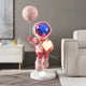 The Stellar Pink Astronaut Glow Heart Balloon Sculpture - 110cm by Giant Sculptures features a pink astronaut with a reflective blue visor, holding a glowing heart and pink balloon. It brings cosmic charm to any modern room, complemented by a white cabinet and gray sofa for an otherworldly yet stylish look.