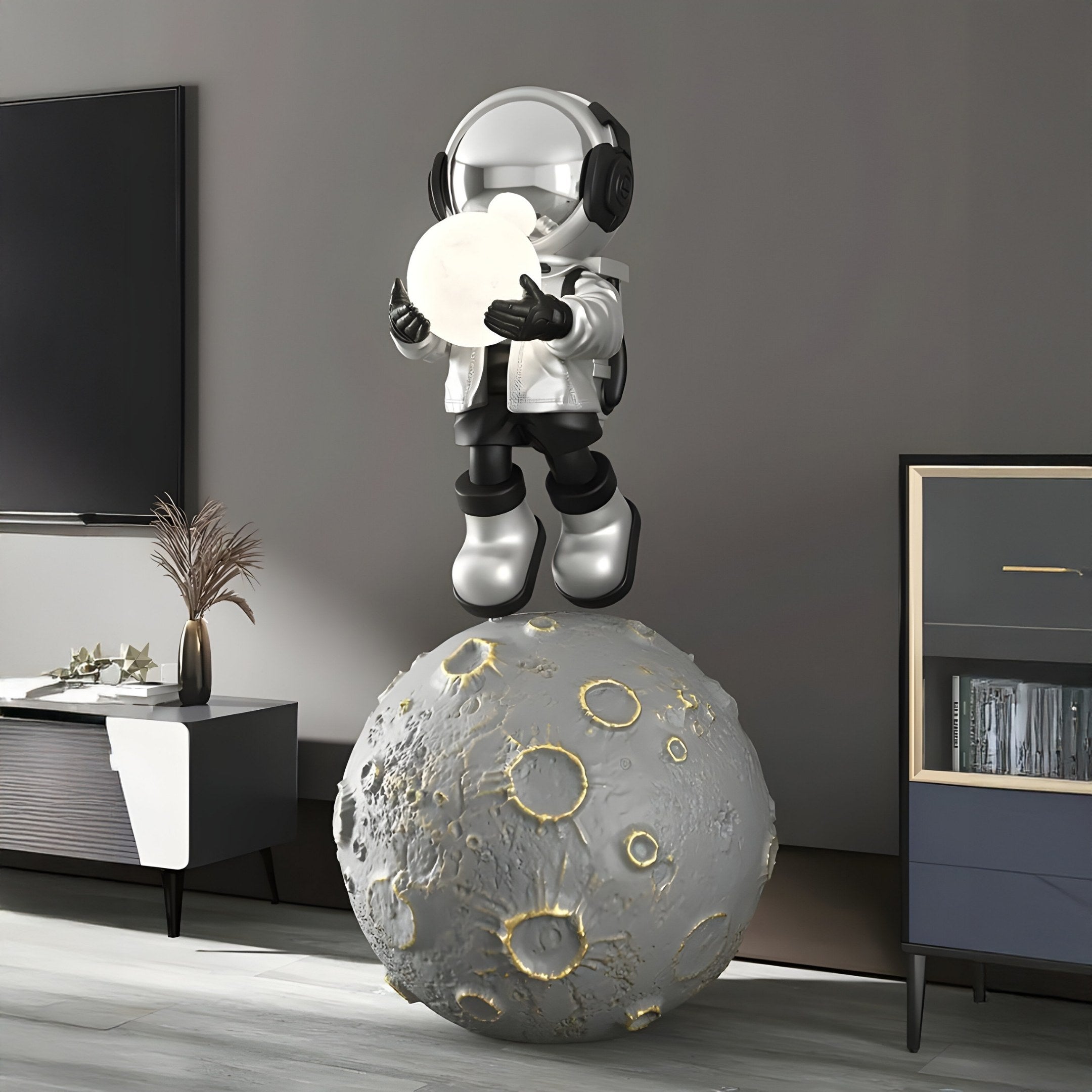 A Lunar Silver Astronaut Glow Moonwalk Sculpture by Giant Sculptures features an astronaut in a white suit and black boots standing on a cratered moon ball, holding a textured moon disc. Its ideal for indoor settings with modern furniture like cabinets and sideboards. Dimensions: 110cm.