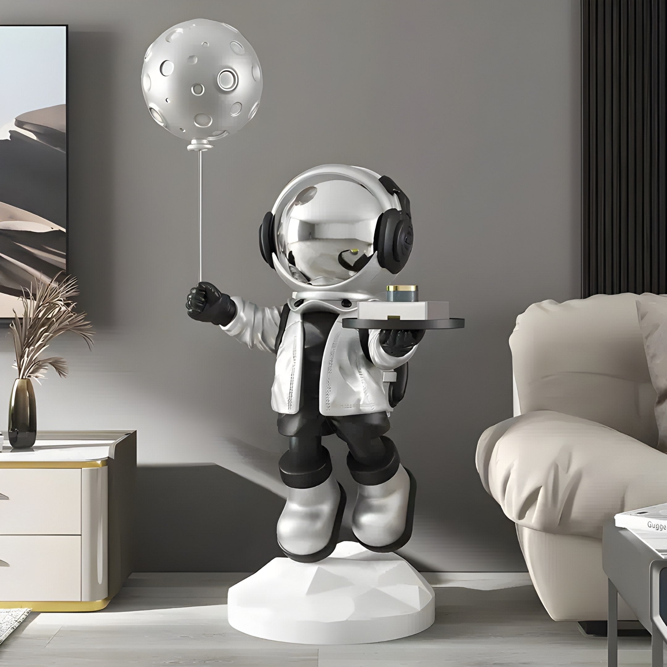 In a modern room, the Lunar Silver Astronaut Butler Balloon Sculpture by Giant Sculptures stands on a white pedestal, holding a moon-like balloon and a tray. Surrounded by neutral tones and minimalistic furniture, it seamlessly blends art with space exploration.