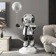 In a modern room, the Lunar Silver Astronaut Butler Balloon Sculpture by Giant Sculptures stands on a white pedestal, holding a moon-like balloon and a tray. Surrounded by neutral tones and minimalistic furniture, it seamlessly blends art with space exploration.