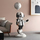 The 110cm Lunar Silver Astronaut Moon Balloon Sculpture by Giant Sculptures, in a sleek spacesuit holding a moon balloon, stands on a textured base in a modern living room with an elegant space-inspired theme, featuring a couch and cat artwork in the background.