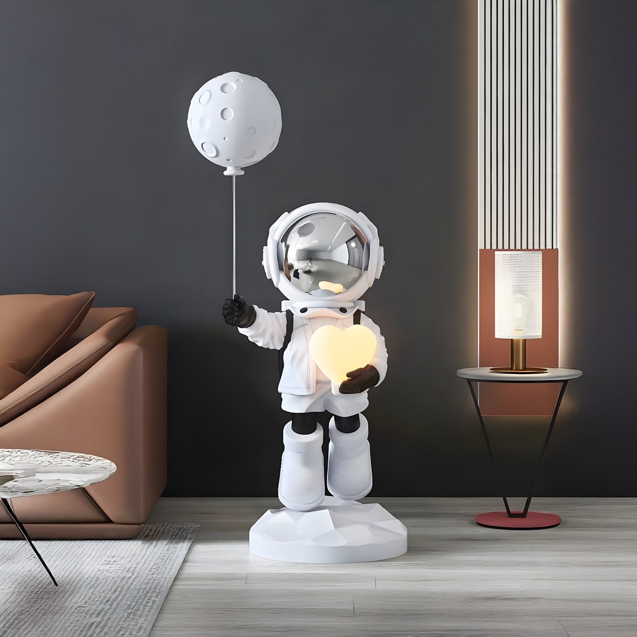 A Nova White Astronaut Glow Heart Balloon Sculpture by Giant Sculptures, featuring an astronaut with a moon-shaped balloon and heart light, stands beside a brown sofa. Nearby is a modern lamp on a stand against a dark vertically lined wall.