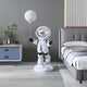 A Nova White Astronaut Moon Balloon Sculpture by Giant Sculptures stands in a modern bedroom with a blue side table holding plants and books next to a gray bed with white bedding, featuring minimalist decor and a striped wall accent.