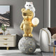 The Gold Astronaut CosmoPals Glow Bear Sculpture by Giant Sculptures features a golden bear standing on a moon-like sphere with a glowing orb, topped by a serene white cat; it includes celestial decor like a modern lamp and white armchair with brown cushion.