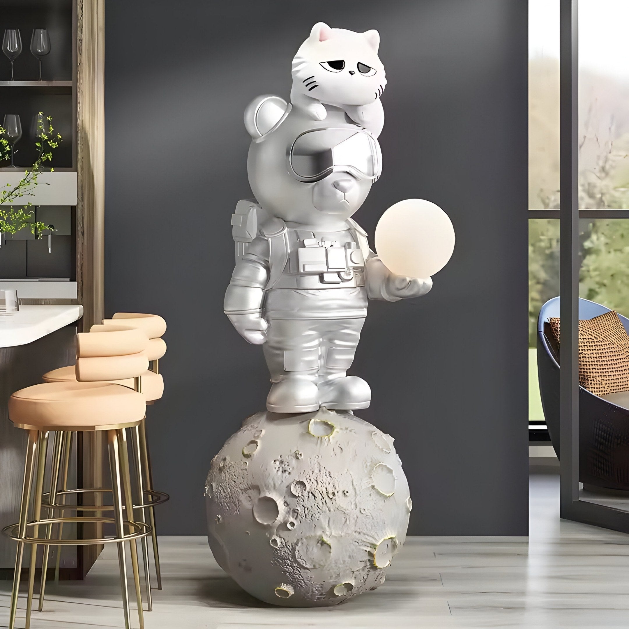 The 115cm Chrome Astronaut CosmoPals Glow Bear Sculpture by Giant Sculptures features a whimsical bear in a chrome astronaut suit on a moon base, holding an orb. A cat perches atop its head against a modern interior with bar stools and expansive views. Ideal for CosmoPals fans!.