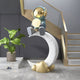 The Galactic Blue Astronaut Glow Moon Crescent Sculpture (116cm) by Giant Sculptures features a gold-headed astronaut on a glowing white crescent moon with gold cloud accents and a yellow sphere, set against gray walls, stairs with gold trim, and a potted plant.