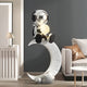 Lunar Silver Astronaut Glow Moon Crescent Sculpture by Giant Sculptures, 116cm resin astronaut with reflective silver helmet seated on a white crescent moon holding a glowing orb, shown in a grey-walled living room beside a sofa.