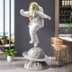 White Space Astronaut Ringed Moon Strider Sculpture by Giant Sculptures, 118cm resin figure in white suit with gold visor mid-stride atop a cratered ringed moon base, shown in a modern bar area.