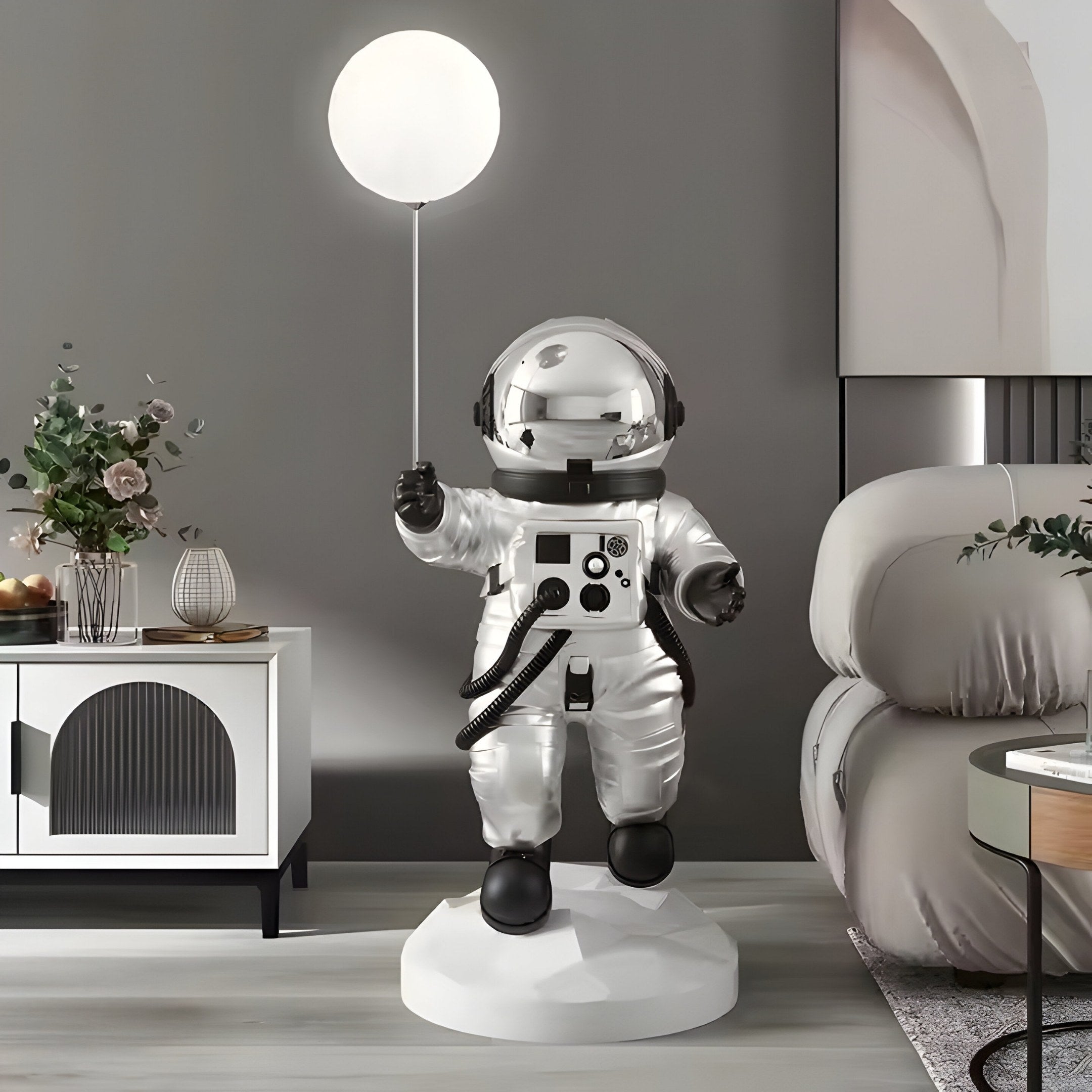 The Lunar Silver Astronaut Ranger Glow Balloon Sculpture by Giant Sculptures stands 120cm tall, depicting an astronaut in a shiny suit holding a glowing balloon. Its set in a modern living room with a beige armchair, decorative cabinet with plants, and a gray wall.