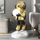 In a modern space, the Celestial Gold Astronaut Glow Hover Sculpture by Giant Sculptures features a reflective helmet and glowing sphere. It stands on a geometric white base beside contemporary furniture like a grey side table and couch, adding both innovation and style.