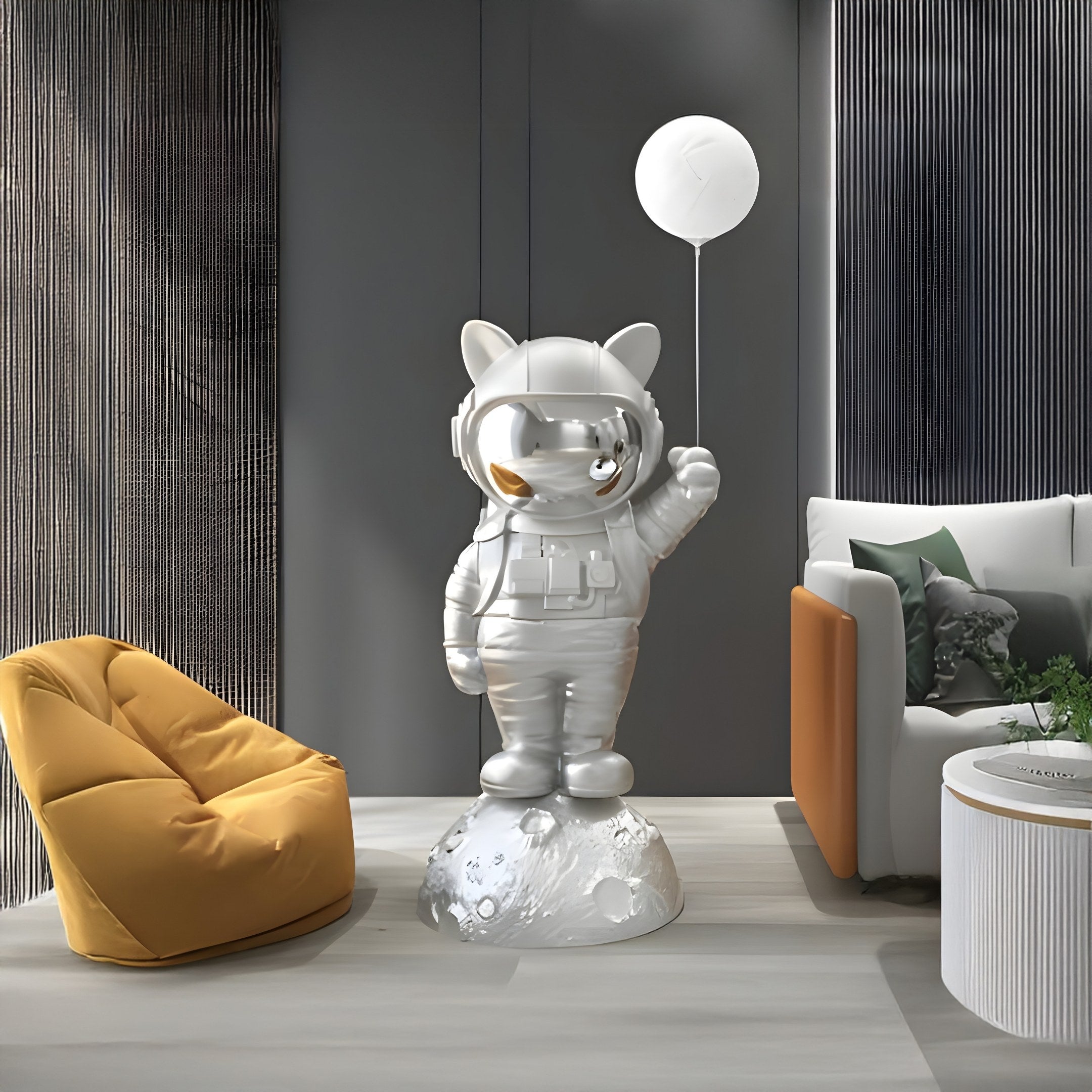 In a modern room, the Cosmic Chrome Astronaut Kitty Glow Balloon Sculpture by Giant Sculptures stands on a moon-like base. A mustard-yellow bean bag chair, gray sofa, and textured gray walls enhance the space-themed décor.