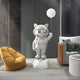 In a modern room, the Cosmic Chrome Astronaut Kitty Glow Balloon Sculpture by Giant Sculptures stands on a moon-like base. A mustard-yellow bean bag chair, gray sofa, and textured gray walls enhance the space-themed décor.