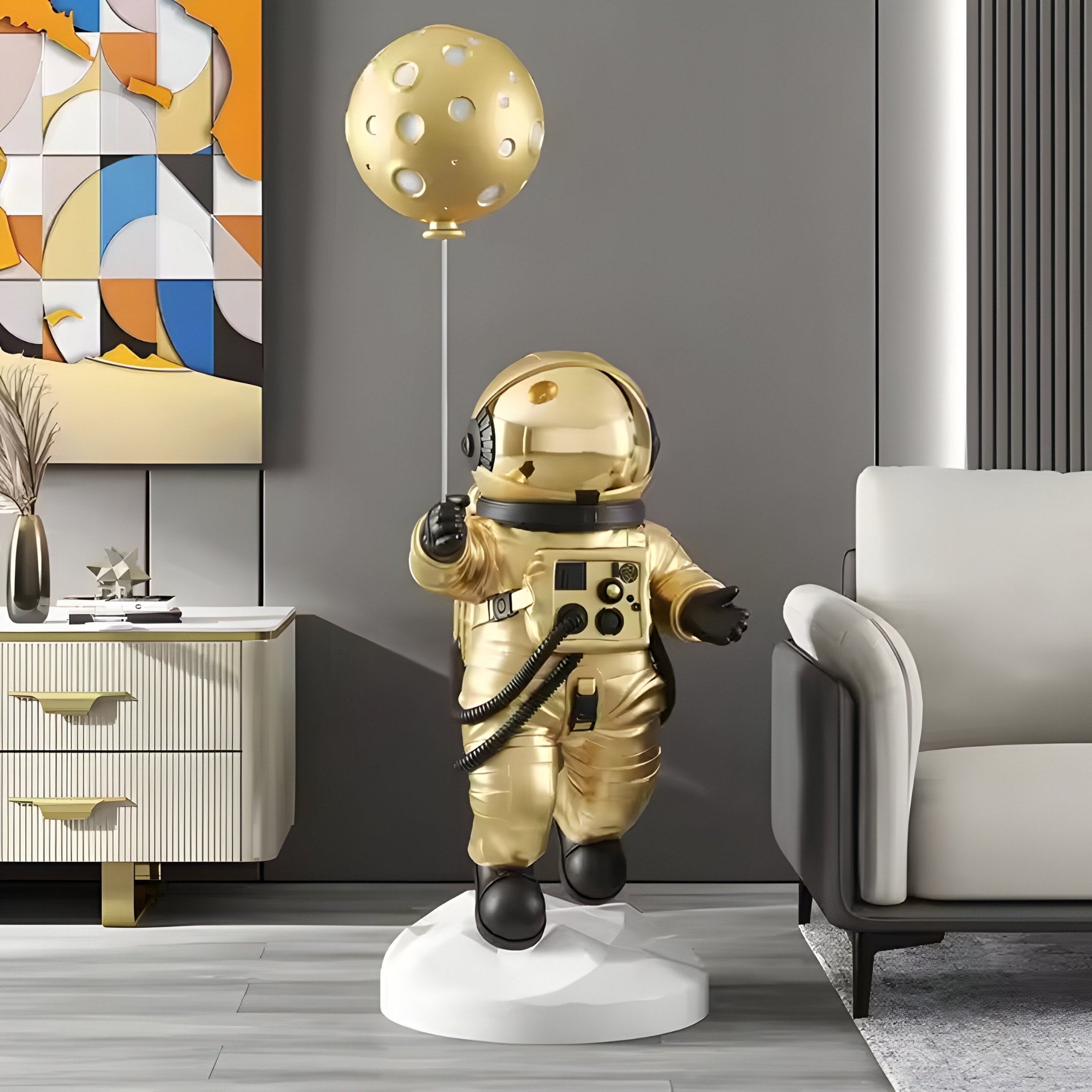 The Celestial Gold Astronaut Ranger Balloon Sculpture by Giant Sculptures, with its shiny gold finish and celestial balloon, stands on a white base. It complements modern decor alongside abstract art, a white sideboard, and an inviting white sofa.