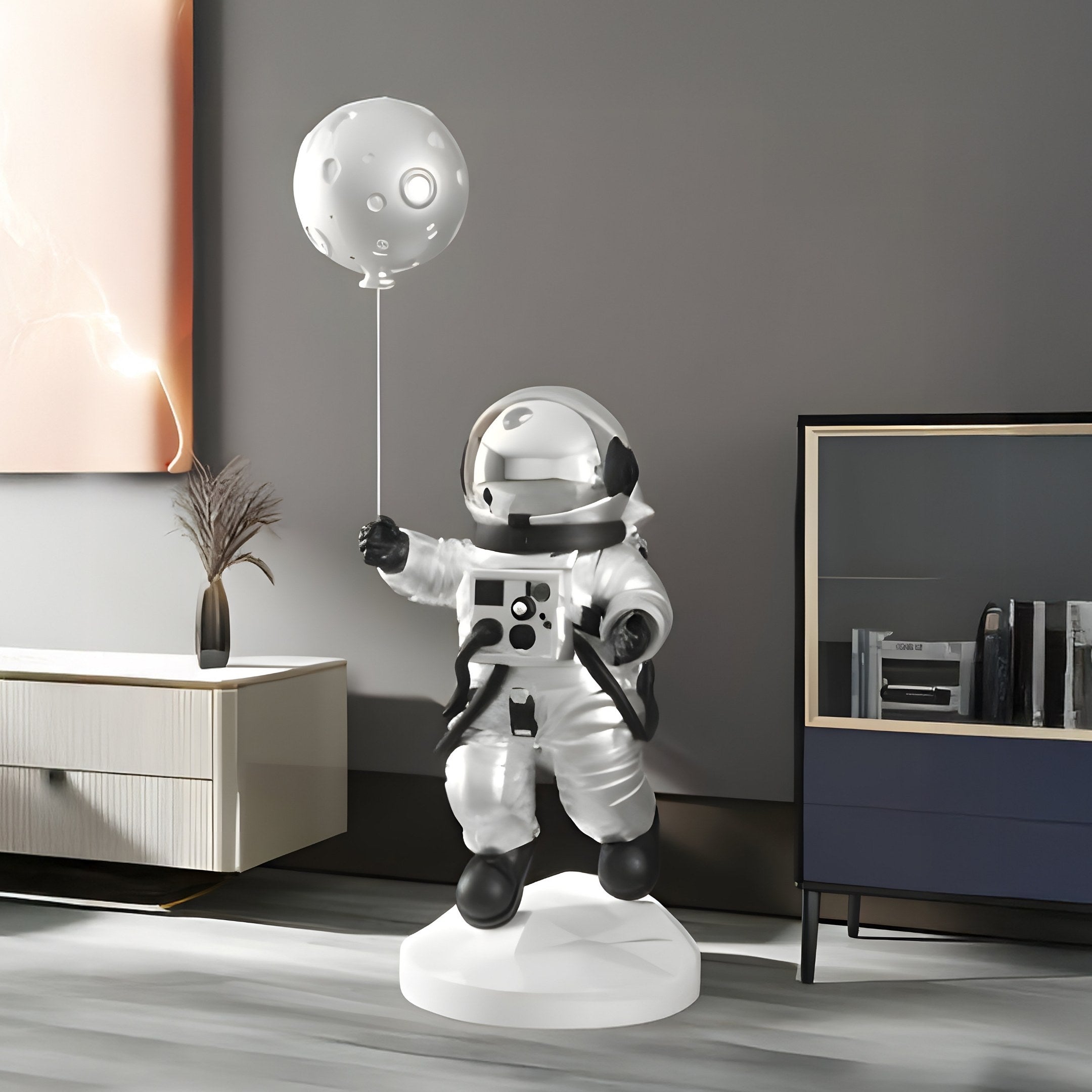 In a modern room with light wood furniture, bookshelves, and a decorative plant, the Lunar Silver Astronaut Ranger Balloon Sculpture - 120cm by Giant Sculptures stands. It depicts an astronaut in a white suit holding a moon-shaped balloon against large abstract wall art embodying space exploration.