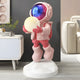 The Stellar Pink Astronaut Glow Hover Sculpture - 120cm by Giant Sculptures showcases a pink astronaut with a blue visor, holding a luminous sphere, on a white base. Perfect for modern living rooms with plants and beige couches, it creates an enchanting ambiance as unique interior decor.