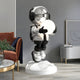 In a modern living room, the Lunar Silver Astronaut Glow Hover Sculpture by Giant Sculptures stands on a white platform, exuding an ethereal glow. Dressed in black and white gear holding a glowing orb, it resembles a touch switch lamp while nearby a brown couch sits adorned with plants and wall art.