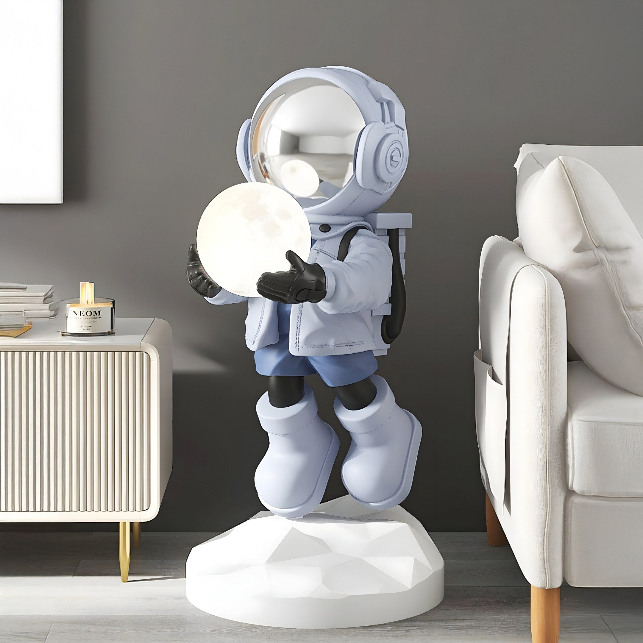 The Giant Sculptures Cosmic Sky Astronaut Glow Hover Sculpture - 120cm features a reflective helmet and large boots on a white base, holding a glowing moon. It stands beside a modern beige sofa and white cabinet with a candle, against a gray wall.