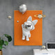 The Giant Sculptures brands Chrome 3D Astronaut Bunny Balloon Orange Wall Art features a resin astronaut sculpture with bunny ears and a planet-shaped balloon floating before an orange board. Nearby, chrome planets hover, while a potted plant and laptop sit on a white desk against the gray wall backdrop.