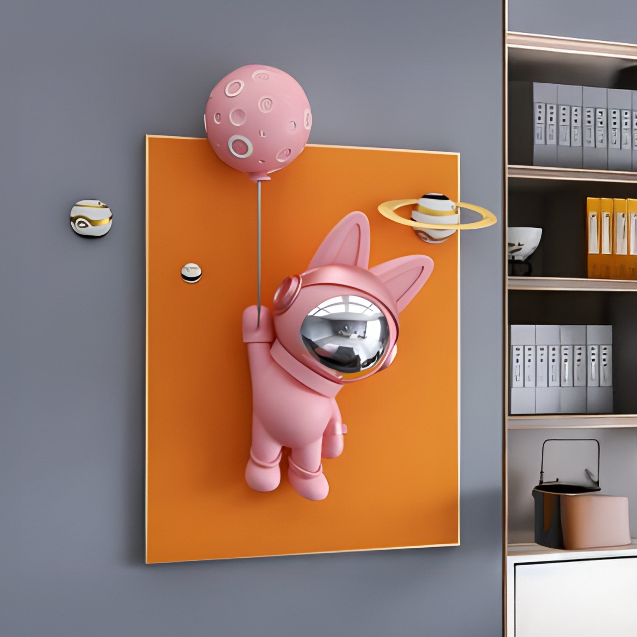 Inspired by the cosmos, the Pink 3D Astronaut Bunny Balloon Orange Wall Art by Giant Sculptures depicts a whimsical bunny with rabbit ears clutching a planet-like balloon against an orange background filled with planets and cars, next to a right-hand bookshelf loaded with books and decor.