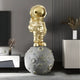 The room features a Celestial Gold Astronaut Star On Moon Sculpture by Giant Sculptures, set on a textured gray sphere. The modern space enthusiast decor includes a dark wall, white cabinet with artful pieces, and a tall white vase with dried plants for a futuristic look.