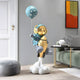 The Giant Sculptures’ Galactic Blue Astronaut Rocket Lift-Off Balloon Sculpture features a blue astronaut with a gold visor and balloon on a cloud base, complemented by stylish decor—a green sofa, gray pouf, and abstract orange-black painting.