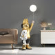 A Gold Astronaut Bunny Pup Glow Balloon Sculpture (130cm) from Giant Sculptures stands amidst space-themed décor, holding a glowing balloon in one hand and a white teddy bear in the other, near a beige sofa and a white cabinet with decorative items.