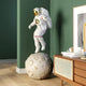 The Giant Sculptures White Space Astronaut Moon Explorer Sculpture (130cm), featuring an astronaut with a gold visor, is poised on a moon-like base. It enhances a modern room with green walls, complemented by a wooden cabinet and framed art.