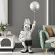 The Silver Astronaut Bunny Pup Balloon Sculpture from Giant Sculptures stands whimsically at 130cm in a modern living room, holding a bear and moon balloon. Its placed near a white console with plants and decor, close to a green sofa, creating an interstellar fantasy scene.