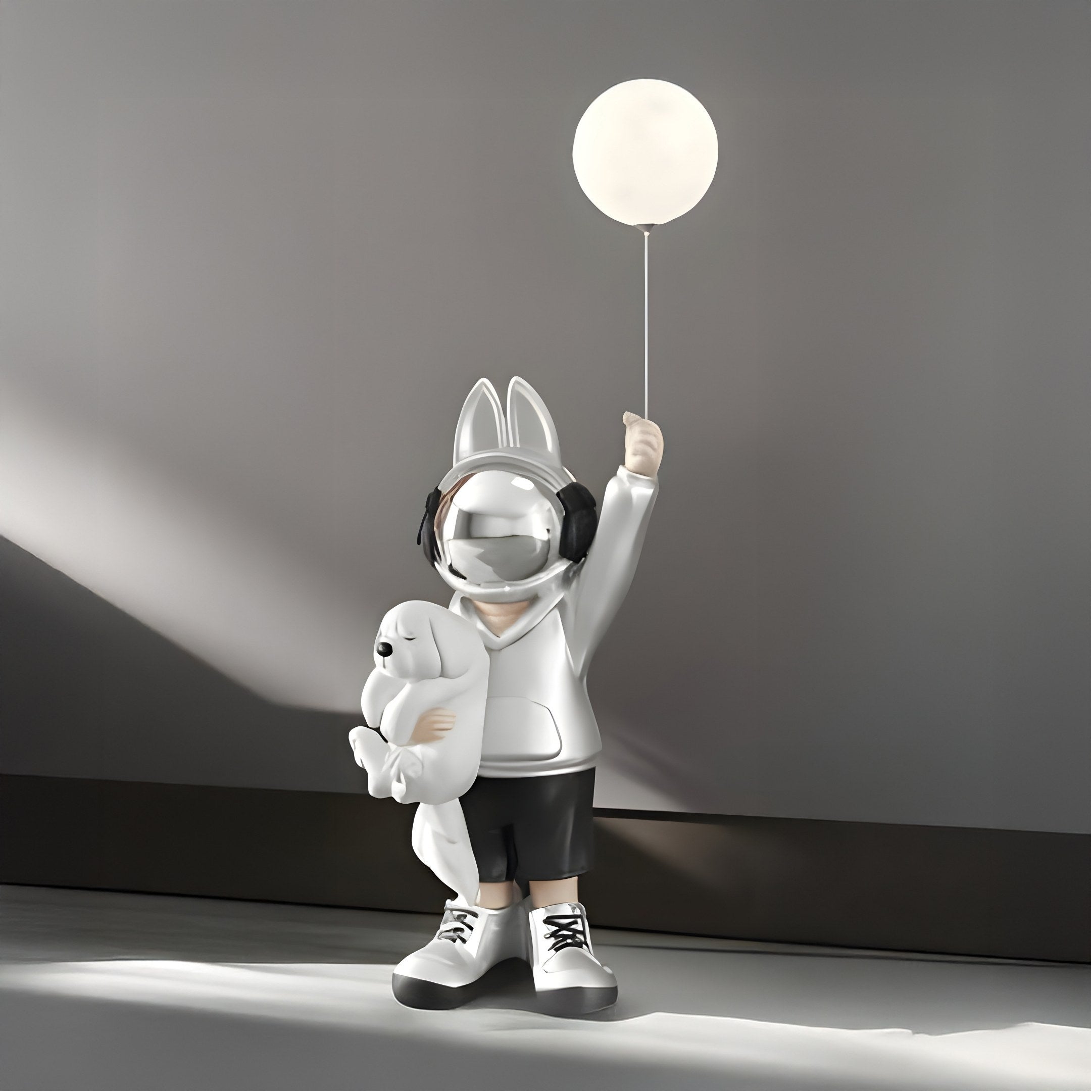 The Silver Astronaut Bunny Pup Glow Balloon Sculpture by Giant Sculptures is a 130cm piece featuring a child in a bunny-eared hoodie with headphones, holding a teddy bear and glow balloon. The sculptures minimalistic background and soft lighting create an enchanting space-themed décor vibe.