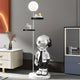 A modern room features the Silver Astronaut Beats Floor Lamp With Shelves - 135cm by Giant Sculptures, enhancing the space-themed décor with its round light and two shelves perfect for glasses or a small plant, alongside potted plants on a cabinet.