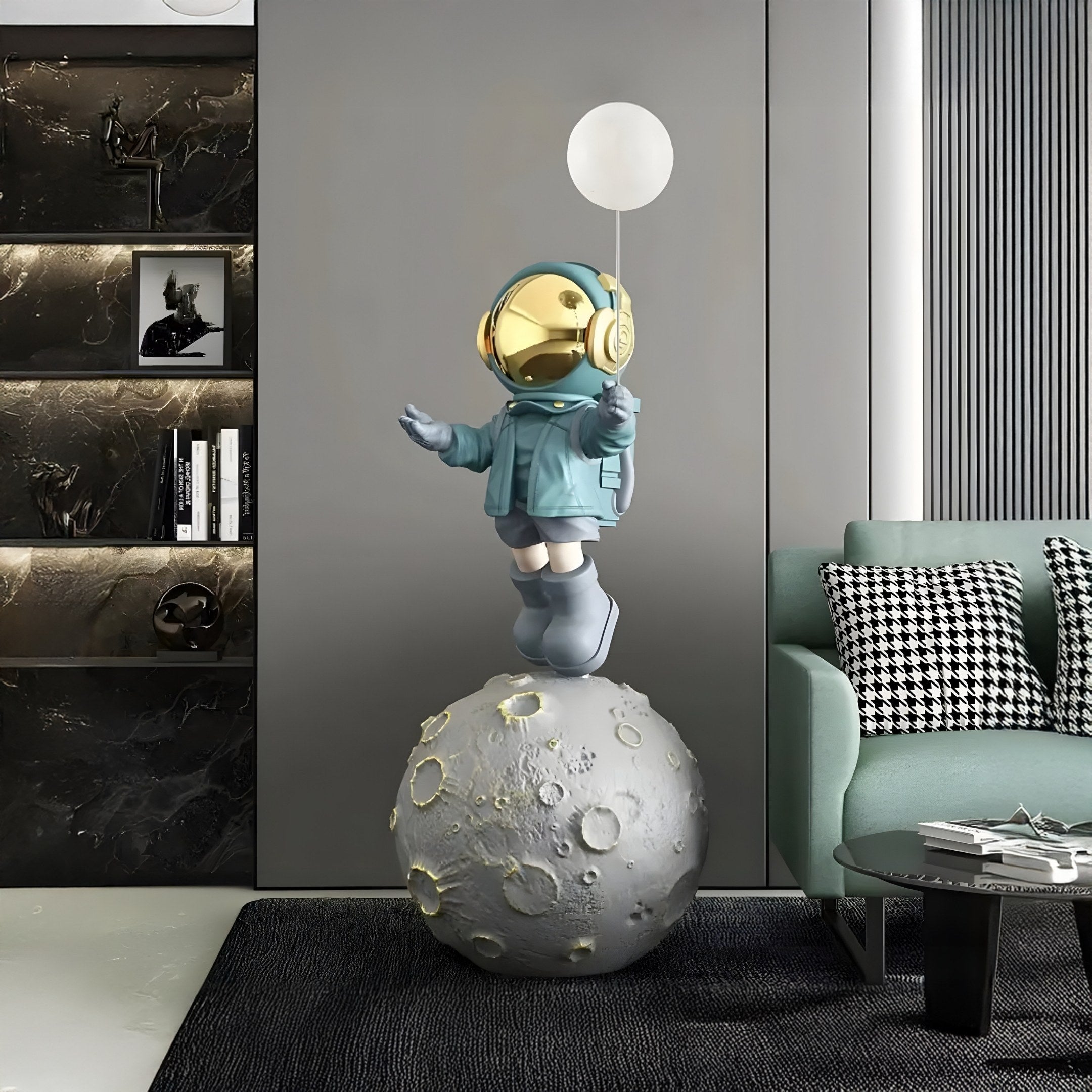 The Galactic Blue Astronaut Glow Balloon Moonwalk Sculpture (140cm) by Giant Sculptures features a futuristic figure with a gold helmet atop a moon-textured sphere, set in a modern living room with dark walls, bookshelf, and gray sofa.