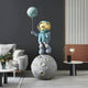 A 140cm Galactic Blue Astronaut Balloon Moonwalk Sculpture by Giant Sculptures is displayed in a modern living room with a sofa, cushions, a floral-adorned round coffee table, and a unique table lamp on a side table against striped wall decor.