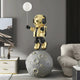 The Celestial Gold Astronaut Glow Balloon Moonwalk Sculpture by Giant Sculptures, standing at 140cm, enhances a modern living room with its cosmic design, featuring an astronaut on a moon sphere holding a glow balloon. Set against neutral tones and abstract art, it charms the plush couch and decor.