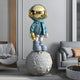 The Galactic Blue Astronaut Beats On Moon Sculpture by Giant Sculptures, featuring a gold helmet and a textured grey lunar sphere, enhances the modern ambiance of a living room with its artistic presence alongside a grey sofa and stylish decorations.