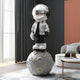 In this modern living room, the Lunar Silver Astronaut Beats On Moon Sculpture - 140cm by Giant Sculptures adds a futuristic touch. The shiny helmet and jacket enhance the décor alongside a small round table with books, a sofa, and a large window accentuating the scene.