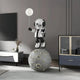 The 140cm Lunar Silver Astronaut Glow Balloon Moonwalk Sculpture by Giant Sculptures elegantly stands on a cratered moon, holding a glowing balloon in a modern room with gray walls, a black cabinet with glass display, minimalist decor, TV, and small vase with dried flowers.