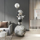 The Lunar Silver Astronaut Balloon Moonwalk Sculpture by Giant Sculptures elevates a space-themed modern living room with its 140cm presence, featuring an astronaut holding a moon-themed balloon atop a large sphere, complementing gray walls, a white sofa with cushions, and sleek wooden floors.