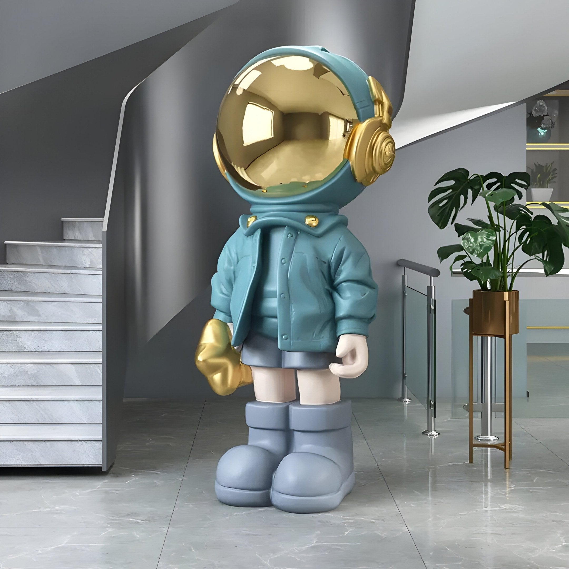 The Galactic Blue Astronaut Beats Sculpture by Giant Sculptures stands 150cm tall indoors beside a staircase, wearing a teal outfit with an oversized jacket, shorts, boots, and a shiny gold reflective helmet. A potted plant is visible in the background, adding to the cosmic decor.