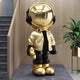 A Celestial Gold Astronaut Beats Sculpture by Giant Sculptures, standing 150cm tall, features a reflective helmet and jacket with black gloves and shoes. Positioned in a modern interior, its flanked by a staircase on the left and a green potted plant on the right, enhancing its cosmic allure.