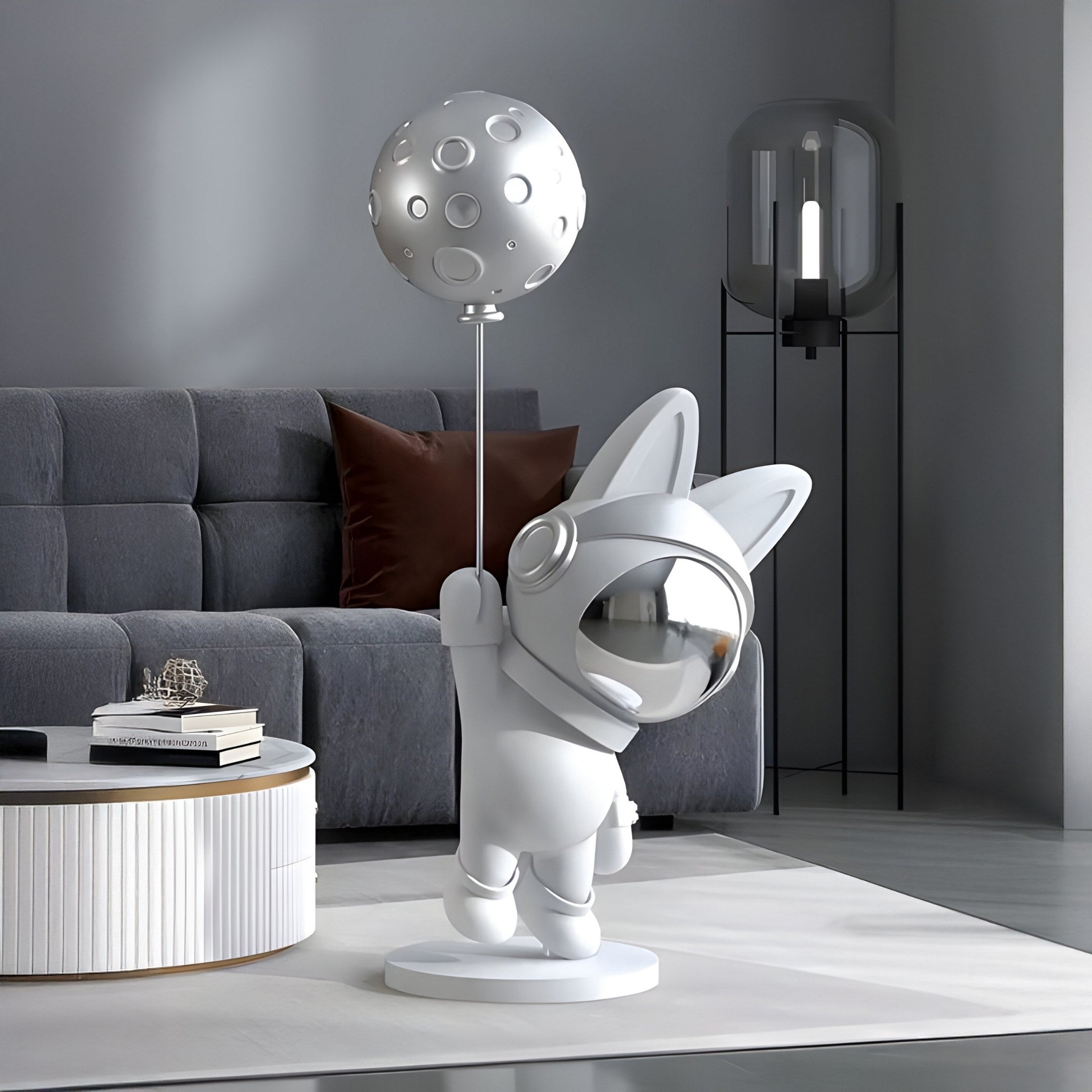 A modern, minimalist room features Giant Sculptures Cosmic Chrome Astronaut Bunny Moon Balloon Sculpture - 150cm. The decor includes a grey sofa, a brown pillow, a round white table with books, and a tall lamp in the background.