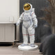 In a modern room, the 150cm White Space Astronaut Standing Sculpture by Giant Sculptures, featuring a reflective gold visor, stands on a sleek white base. It epitomizes space exploration decor and is surrounded by minimalistic furniture like a potted plant and an understated cabinet.