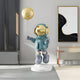 The Galactic Blue Astronaut Moon Balloon Sculpture - 160cm by Giant Sculptures features a space-themed figure in a shiny helmet and blue outfit floating above a stand. Enhance your modern decor with its cosmic energy alongside a black and white abstract painting and sleek wooden console table.