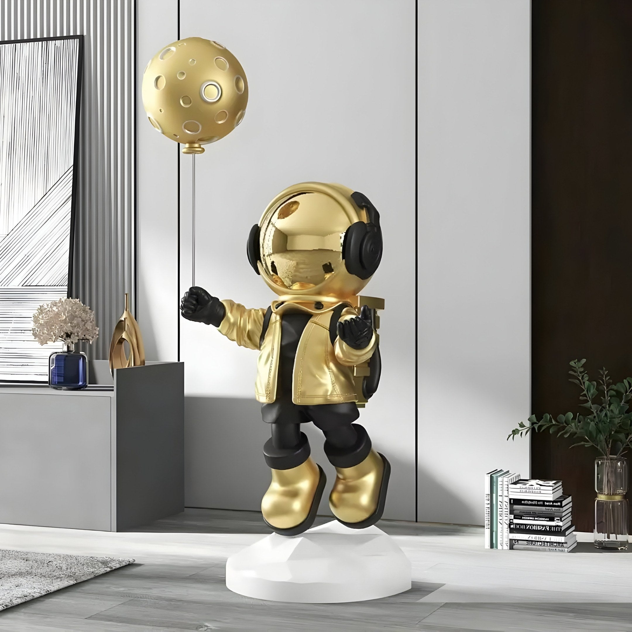 The Celestial Gold Astronaut Moon Balloon Sculpture from Giant Sculptures features a gold and black astronaut with headphones, standing on a white base, holding a moon balloon. This 160cm sculpture complements modern decor with plants, books, and art for a sleek futuristic vibe.