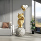 In a modern room with a white sofa, brown cushions, and a potted plant by tall windows, natural light creates serenity as the Gilded Gold Astronaut Kitty Heart Balloons Sculpture by Giant Sculptures stands on a lunar sphere holding silver heart-shaped balloons.