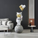 A modern living room features Giant Sculptures Nova White Astronaut Kitty Heart Balloons Sculpture, 160cm tall. The astronaut has a gold visor and heart-shaped balloons, standing on a moon-like base. A white sofa pairs with a round coffee table and side table with a lamp for a cosmic vibe.